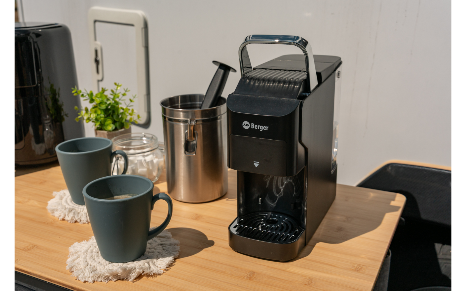 Zwarte capsulekoffiemachine op houten aanrecht, twee donkere mokken op onderzetters, roestvrijstalen bak met capsuleschep, glazen pot met deksel, kleine potplant