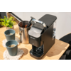 Zwarte koffiemachine op een licht houten tafel, twee grijze keramische kopjes op gehaakte onderzetters, geopend metalen blik met houten schep, kleine groene kamerplant op de achtergrond, stoelrug aan rechts