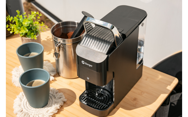 Zwarte koffiemachine op een licht houten tafel, twee grijze keramische kopjes op gehaakte onderzetters, geopend metalen blik met houten schep, kleine groene kamerplant op de achtergrond, stoelrug aan rechts