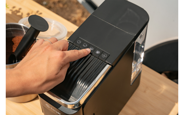 Hand drukt bedieningspaneel van een zwarte koffiemachine op een houten oppervlak; daarnaast een bakje met koffiepoeder en een zwarte maatschep