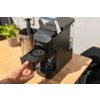 Hand trekt een capsulelade uit een zwarte koffiemachine op een houten werkblad; melkkan en kleine potplant op de achtergrond