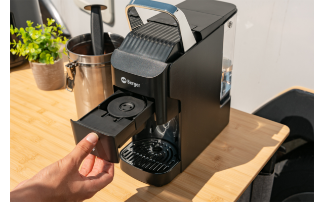 Hand trekt een capsulelade uit een zwarte koffiemachine op een houten werkblad; melkkan en kleine potplant op de achtergrond