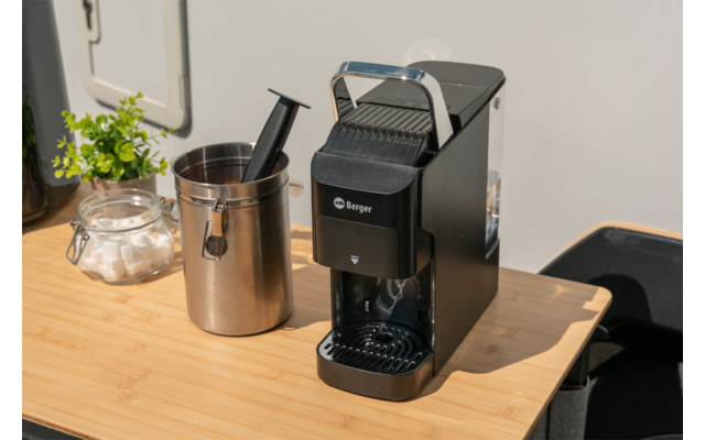 Zwart koffieapparaat op licht houten tafel, daarnaast een roestvrijstalen melkkannetje, glazen pot met suikerklontjes, kleine kamerplant en een stoel