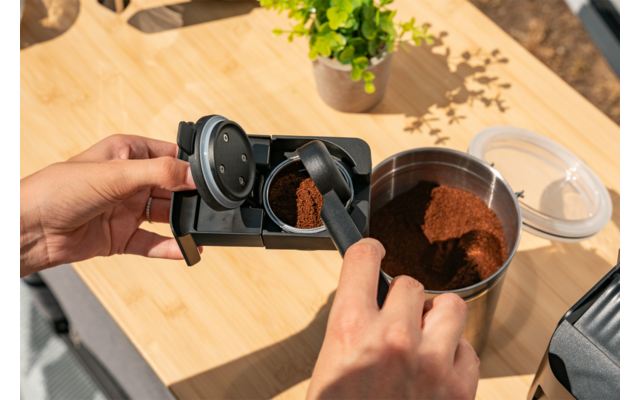 Twee handen scheppen gemalen koffie met een zwarte lepel in een metalen portafilter op een houten oppervlak; een open metalen bakje met koffiepoeder en een kleine potplant zijn zichtbaar