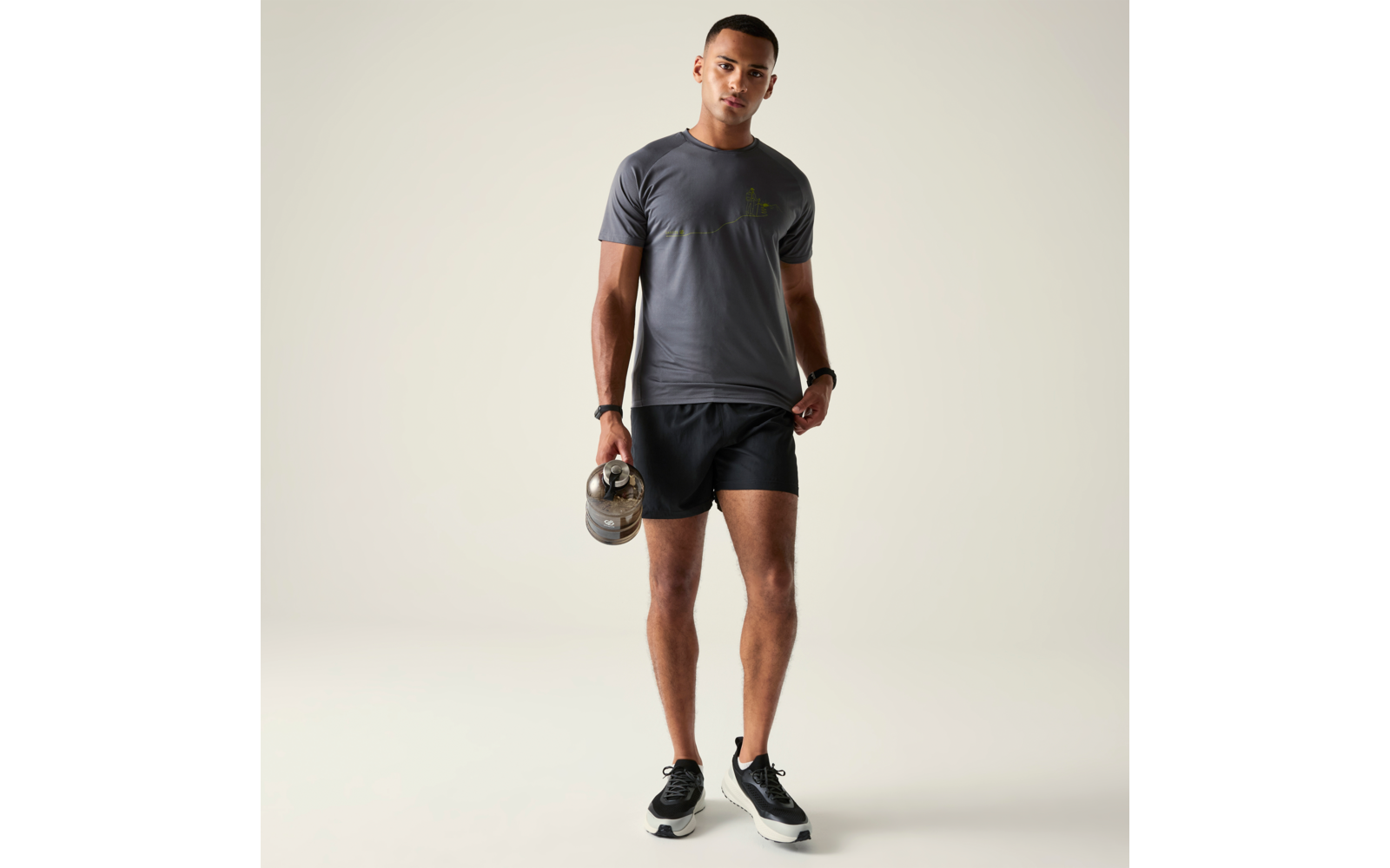 Person steht frontal vor hellem Hintergrund, trägt graues T‑Shirt, schwarze Shorts und schwarze Sportschuhe, hält eine Hantel in der rechten Hand