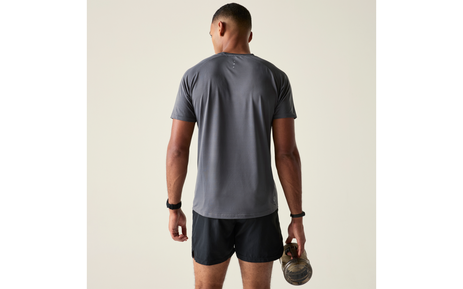 Person von hinten in grauem T-Shirt und schwarzen Shorts, hält eine Kettlebell in der rechten Hand vor beigem Hintergrund