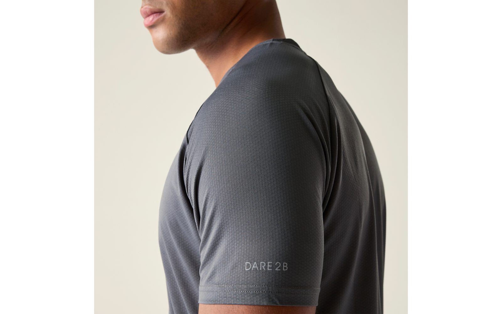 Seitliche Aufnahme des Oberkörpers einer Person mit sichtbarem Kinn und Unterlippe, sie trägt ein dunkelgraues Kurzarmshirt mit dem Schriftzug DARE 2B am Ärmel, heller einfarbiger Hintergrund