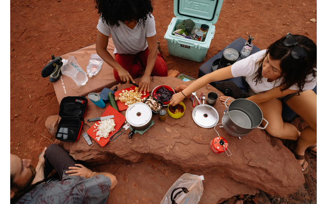 Tre persone sedute su terra rossa attorno a una roccia piatta; sulla roccia fornelli da campeggio, pentole, un tagliere con verdure tritate, ciotole e una ghiacciaia con verdure