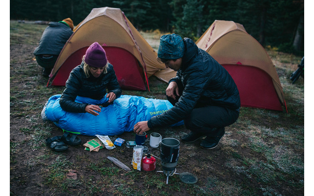 Due persone con abbigliamento da esterno accanto a due tende giallo-rosse; una persona è distesa in un sacco a pelo blu a terra, l'altra è accovacciata accanto; attrezzatura da campeggio (fornello, pentole, confezioni di cibo, sandali) in primo piano; bosco di conifere sullo sfondo