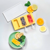 Molde de silicona amarillo para polos con dos polos (azul oscuro, rosa) y una cavidad vacía; encima una tapa transparente. Abajo, un plato verde con un polo amarillo, rodajas de limón, arándanos y hojas de menta. Rodajas de limón y naranja a la derecha y arriba