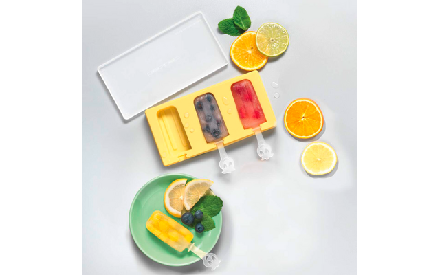 Molde de silicona amarillo para polos con dos polos (azul oscuro, rosa) y una cavidad vacía; encima una tapa transparente. Abajo, un plato verde con un polo amarillo, rodajas de limón, arándanos y hojas de menta. Rodajas de limón y naranja a la derecha y arriba