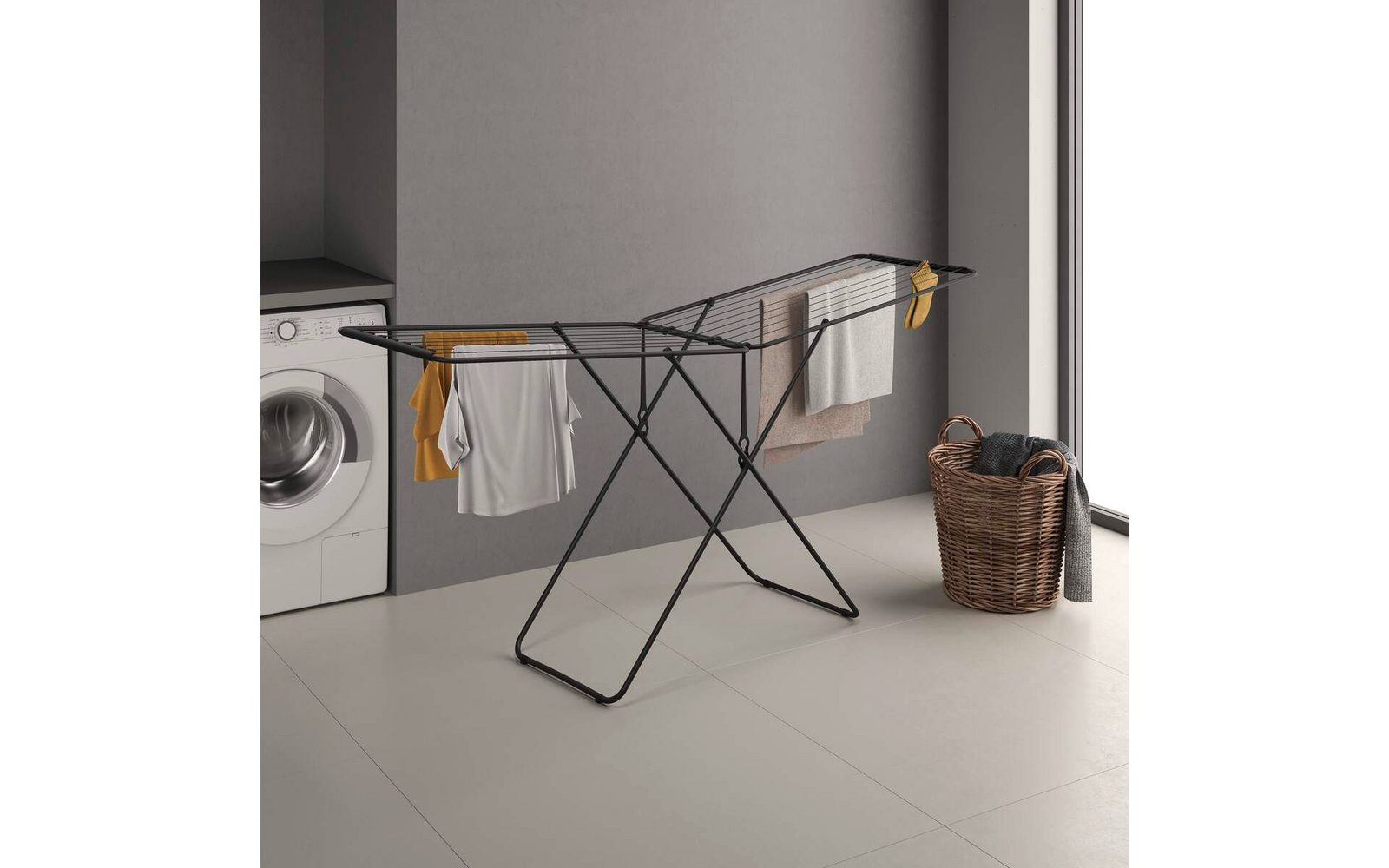 Zwart inklapbaar droogrek met een wit T‑shirt en gele en beige doeken hangend op een lichte betegelde vloer; links een wasmachine, rechts een gevlochten wasmand met kleding