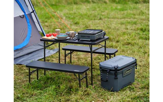 Grill und Geschirr auf einem kleinen schwarzen Tisch im Gras, daneben eine Kühlbox und ein Zelt