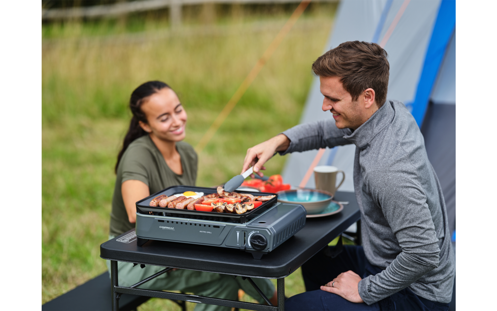Zwei Personen grillen gemeinsam im Freien, eine Frau und ein Mann, der mit einer Zange Würstchen auf dem Grill wendet