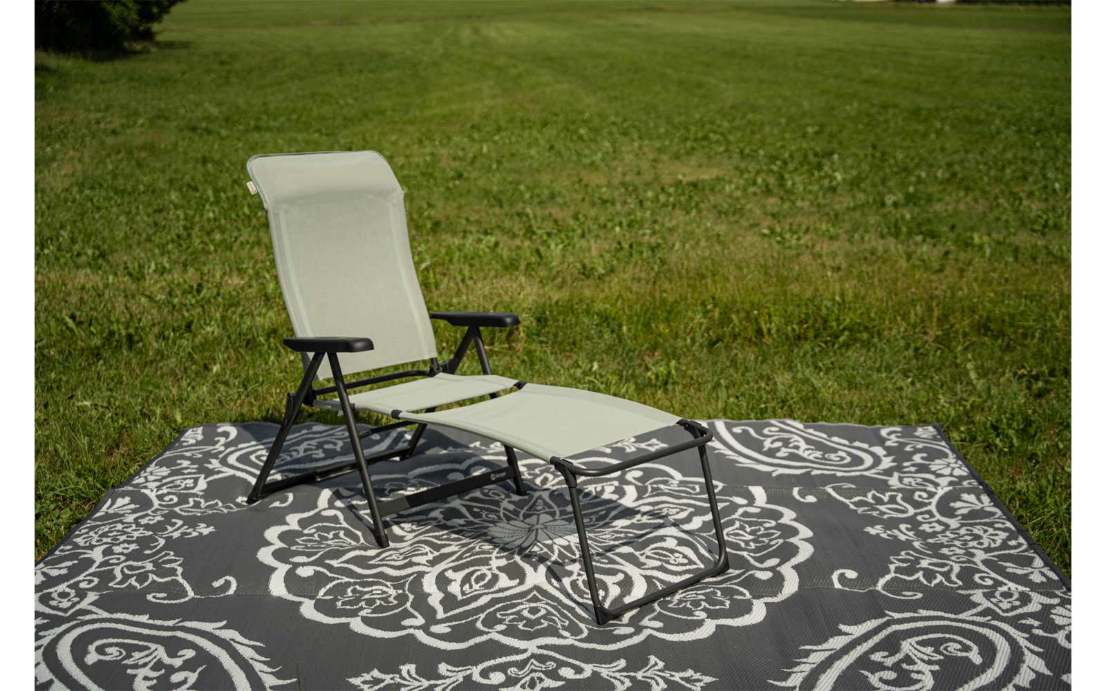 Leichtgrüner Liegestuhl mit schwarzem Metallgestell auf grauem Teppich mit ornamentalem Muster auf einer Grasfläche