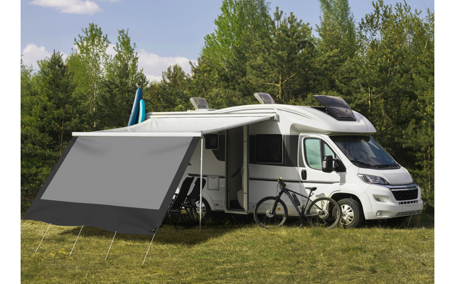 Witte camper op gras met uitgeklapte grijze luifel, twee fietsen naast het voertuig, bomen op de achtergrond en een blauwe lucht met wolken