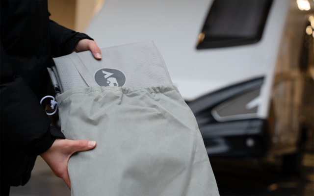 Dos manos sostienen una bolsa gris claro de la que sobresale un cojín plegado gris claro con un logo redondo oscuro; en el fondo se ve un vehículo blanco