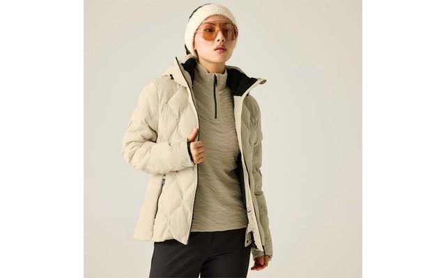 Person trägt beigefarbene Steppjacke, hellen Pullover mit kurzem Reißverschluss, beigefarbene Mütze, Sonnenbrille und dunkle Hosen vor hellem einfarbigen Hintergrund