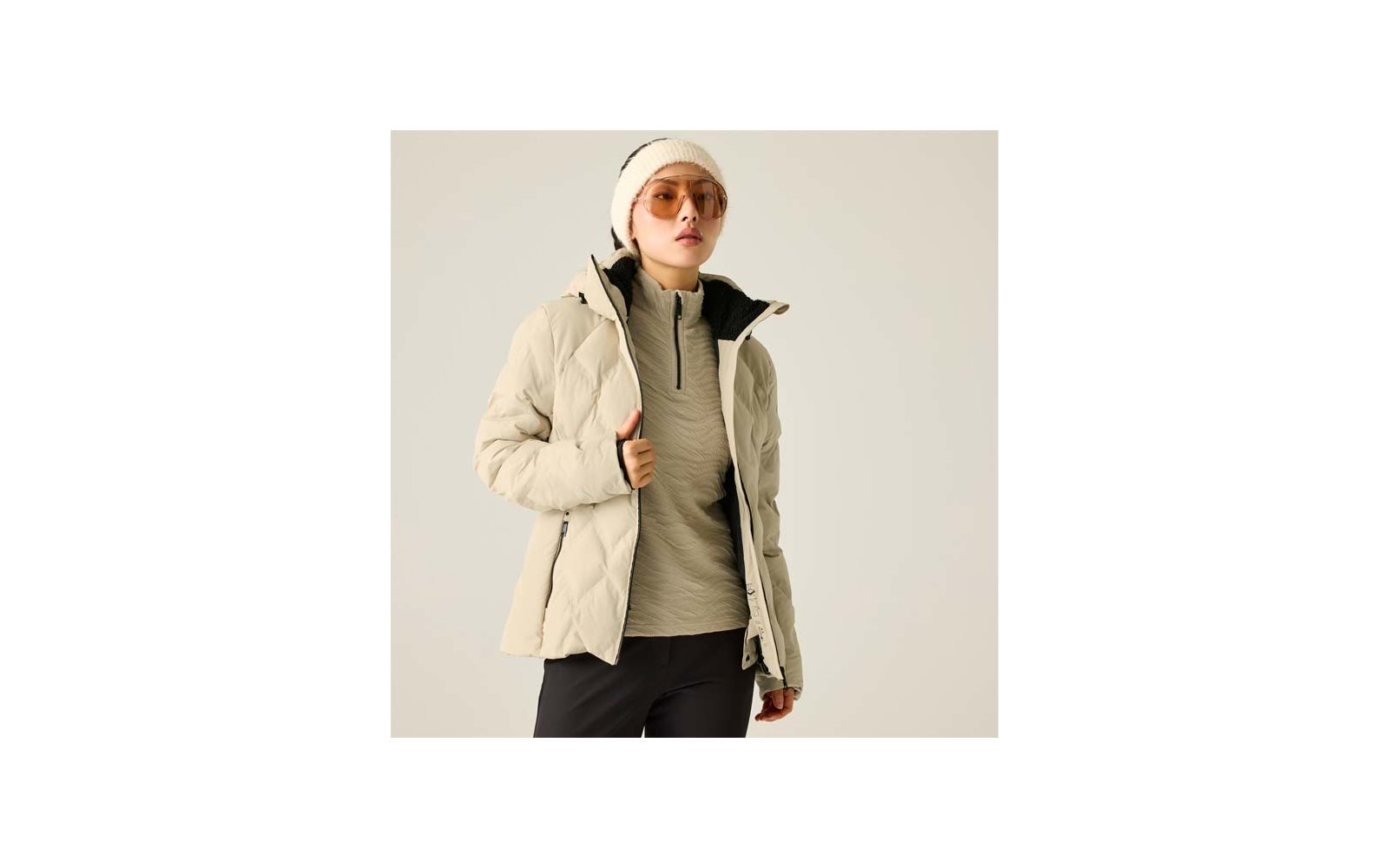 Person trägt beigefarbene Steppjacke, hellen Pullover mit kurzem Reißverschluss, beigefarbene Mütze, Sonnenbrille und dunkle Hosen vor hellem einfarbigen Hintergrund