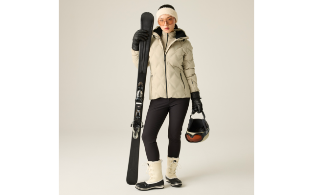 Person steht, hält einen Ski senkrecht in der rechten Hand und einen Skihelm in der linken Hand, trägt beige Winterjacke, schwarze Skihose, weiße Skistiefel, Handschuhe, Stirnband und Skibrille