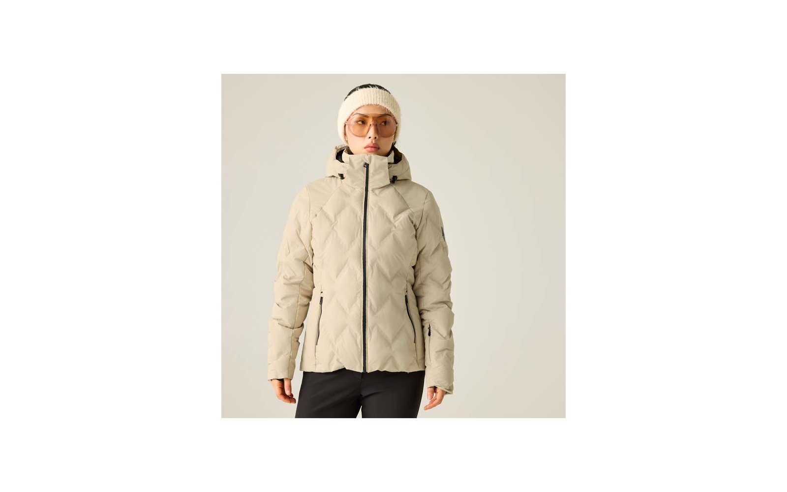 Person vor hellem Hintergrund, weiße Strickmütze, orangegetönte Brille, beige gesteppte Jacke mit Kapuze und Reißverschluss, schwarze Hose