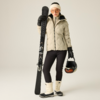Person steht, hält einen Ski senkrecht in der rechten Hand und einen Skihelm in der linken Hand, trägt beige Winterjacke, schwarze Skihose, weiße Skistiefel, Handschuhe, Stirnband und Skibrille