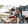 Zwei Personen sitzen an einem Klapptisch vor einem geöffneten Wohnmobil. Auf dem Tisch stehen Kaffeetassen, eine Thermoskanne, eine Kaffeemühle und eine kleine Pflanze. Im Hintergrund ein See, Bäume und eine ausgefahrene Markise