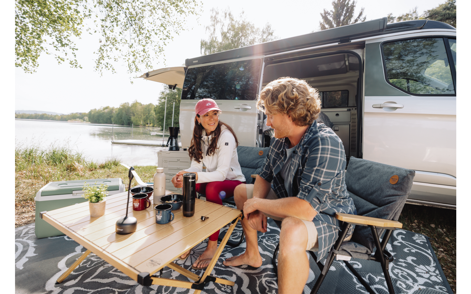 Zwei Personen sitzen an einem Klapptisch vor einem geöffneten Wohnmobil. Auf dem Tisch stehen Kaffeetassen, eine Thermoskanne, eine Kaffeemühle und eine kleine Pflanze. Im Hintergrund ein See, Bäume und eine ausgefahrene Markise
