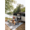 Wohnmobil mit geöffneter Schiebetür am See. Zwei Personen sitzen auf Campingstühlen an einem kleinen Klapptisch auf einem gemusterten Outdoor-Teppich; Becher und eine Kaffeekanne stehen auf dem Tisch. Bäume am Ufer im Hintergrund