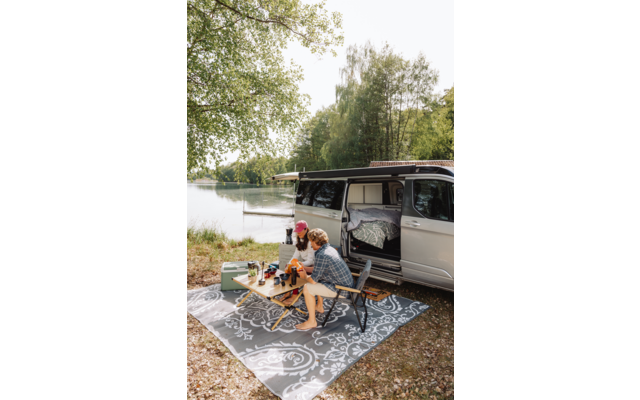 Wohnmobil mit geöffneter Schiebetür am See. Zwei Personen sitzen auf Campingstühlen an einem kleinen Klapptisch auf einem gemusterten Outdoor-Teppich; Becher und eine Kaffeekanne stehen auf dem Tisch. Bäume am Ufer im Hintergrund