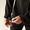 Nahaufnahme von zwei Händen, die einen Klettverschluss am Ärmel einer schwarzen Jacke schließen; Reißverschluss, Oberkörper und schwarze Hose sichtbar