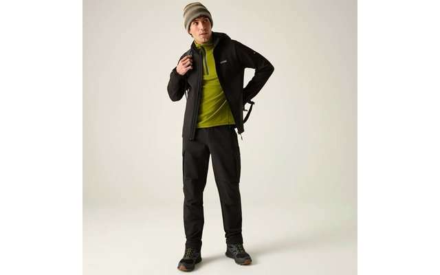 Person steht vor hellem Hintergrund, trägt graue Mütze, grünen Pullover, schwarze Jacke, schwarze Hose und Wanderschuhe