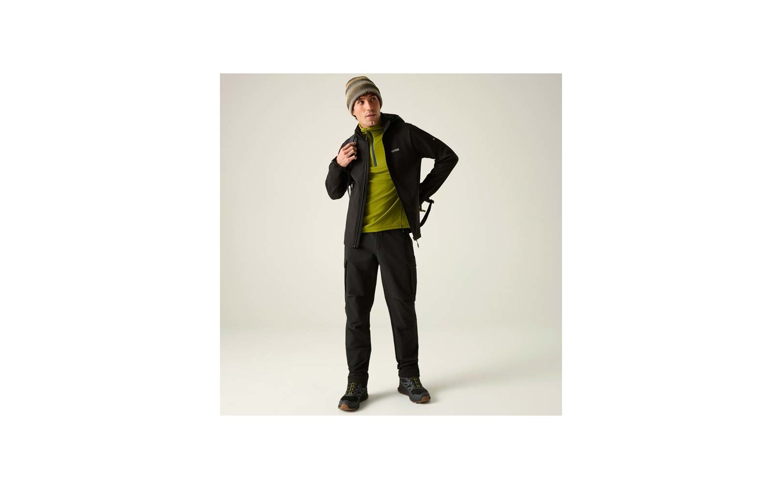 Person steht vor hellem Hintergrund, trägt graue Mütze, grünen Pullover, schwarze Jacke, schwarze Hose und Wanderschuhe