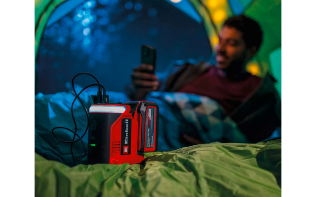 Rotes tragbares Ladegerät mit angeschlossenen Kabeln in einem Zelt; im Hintergrund eine verschwommene Person im Schlafsack, die ein Smartphone hält
