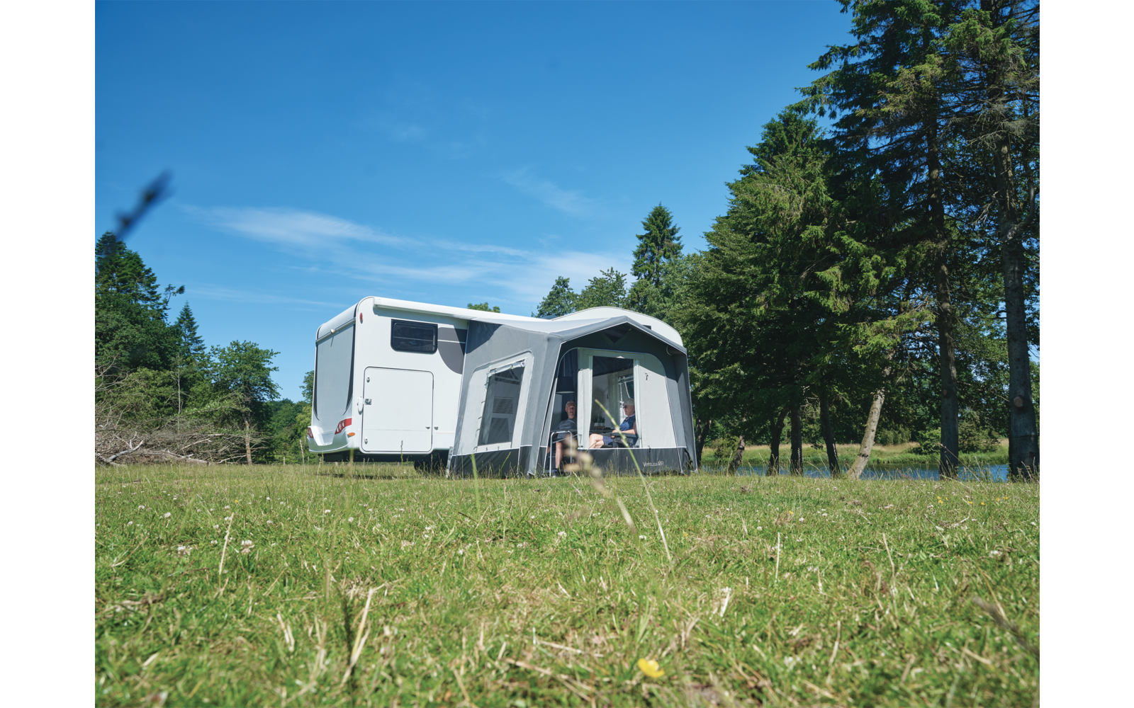 Weißer Wohnwagen mit grauem Vorzelt auf einer Wiese, Bäume im Hintergrund und blauer Himmel