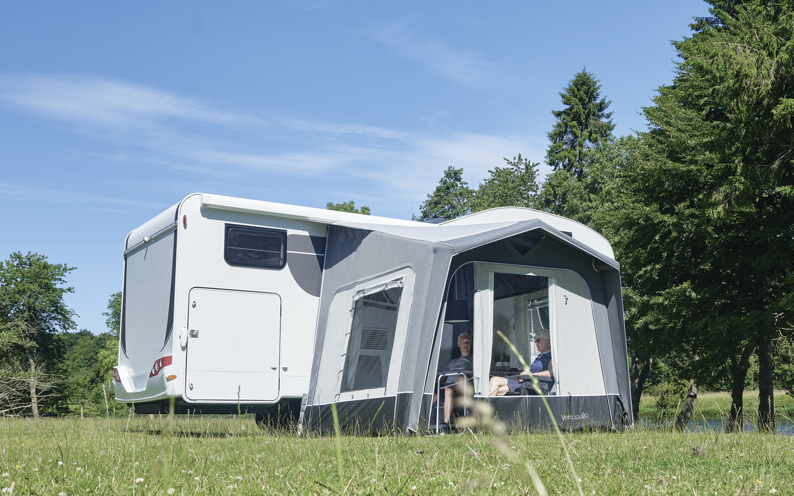 Weißer Wohnwagen mit grauem Vorzelt auf einer Wiese, Bäume im Hintergrund und blauer Himmel
