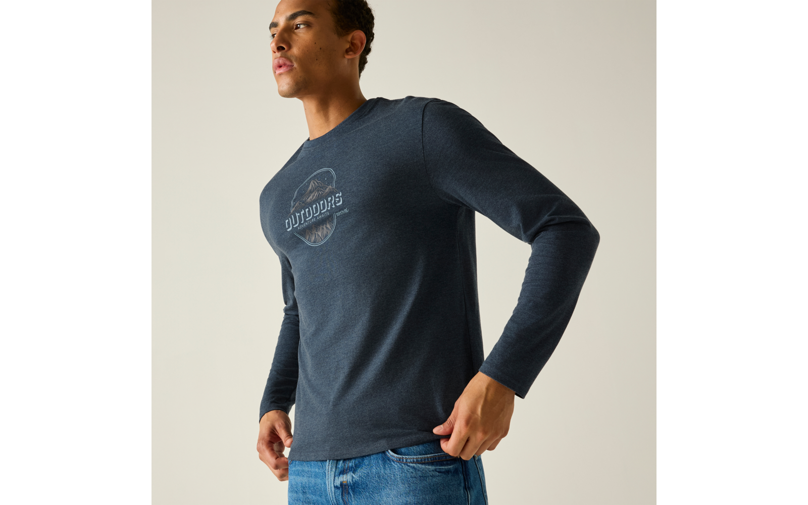 Persoon in driekwartprofiel draagt donkerblauw longsleeve met rond 'OUTDOORS' logo op de borst, blauwe spijkerbroek, handen bij de heupen, effen lichte achtergrond