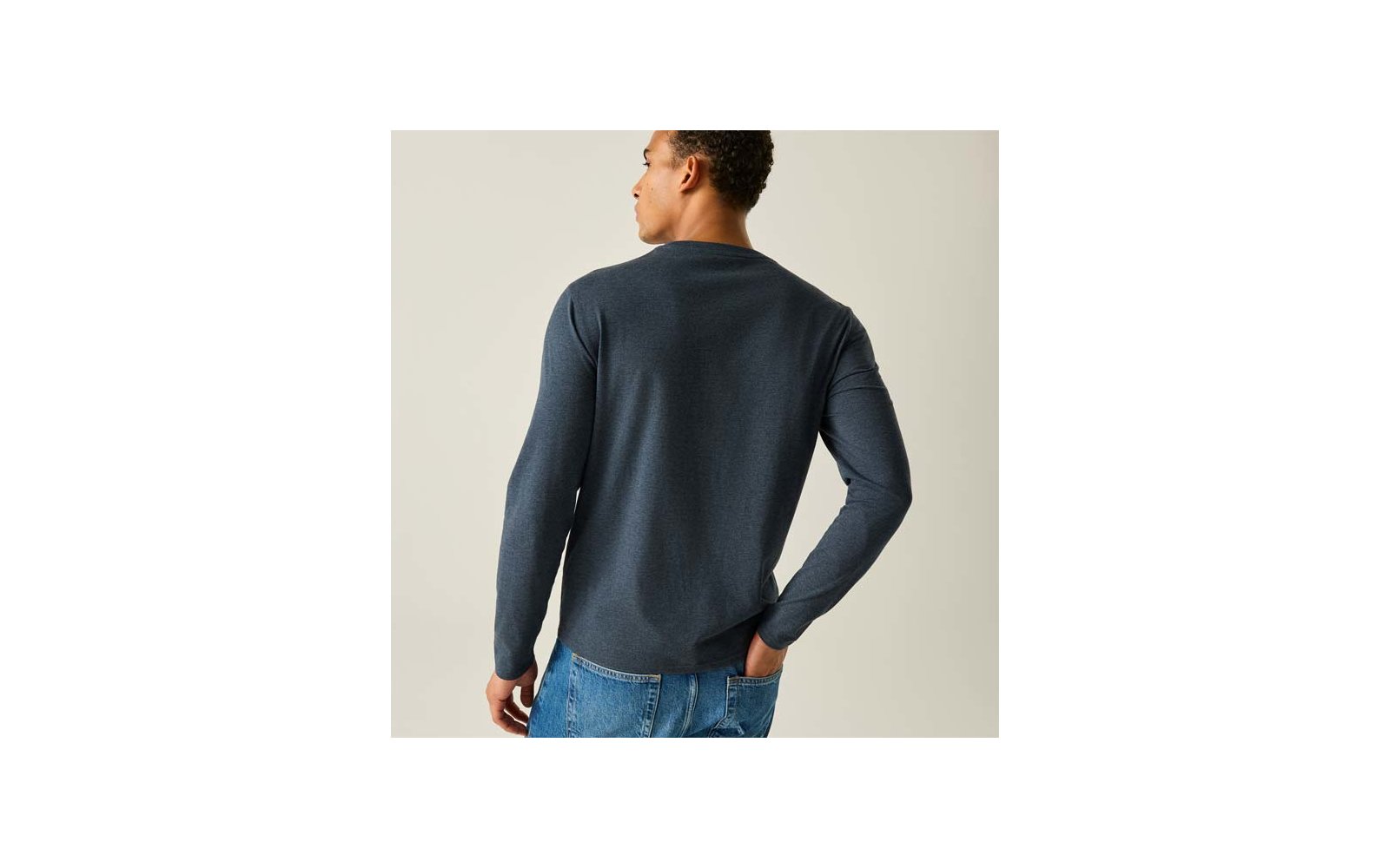 Persoon van achteren, draagt donker shirt met lange mouwen en blauwe spijkerbroek, linkerhand in achterzak, neutrale lichte achtergrond