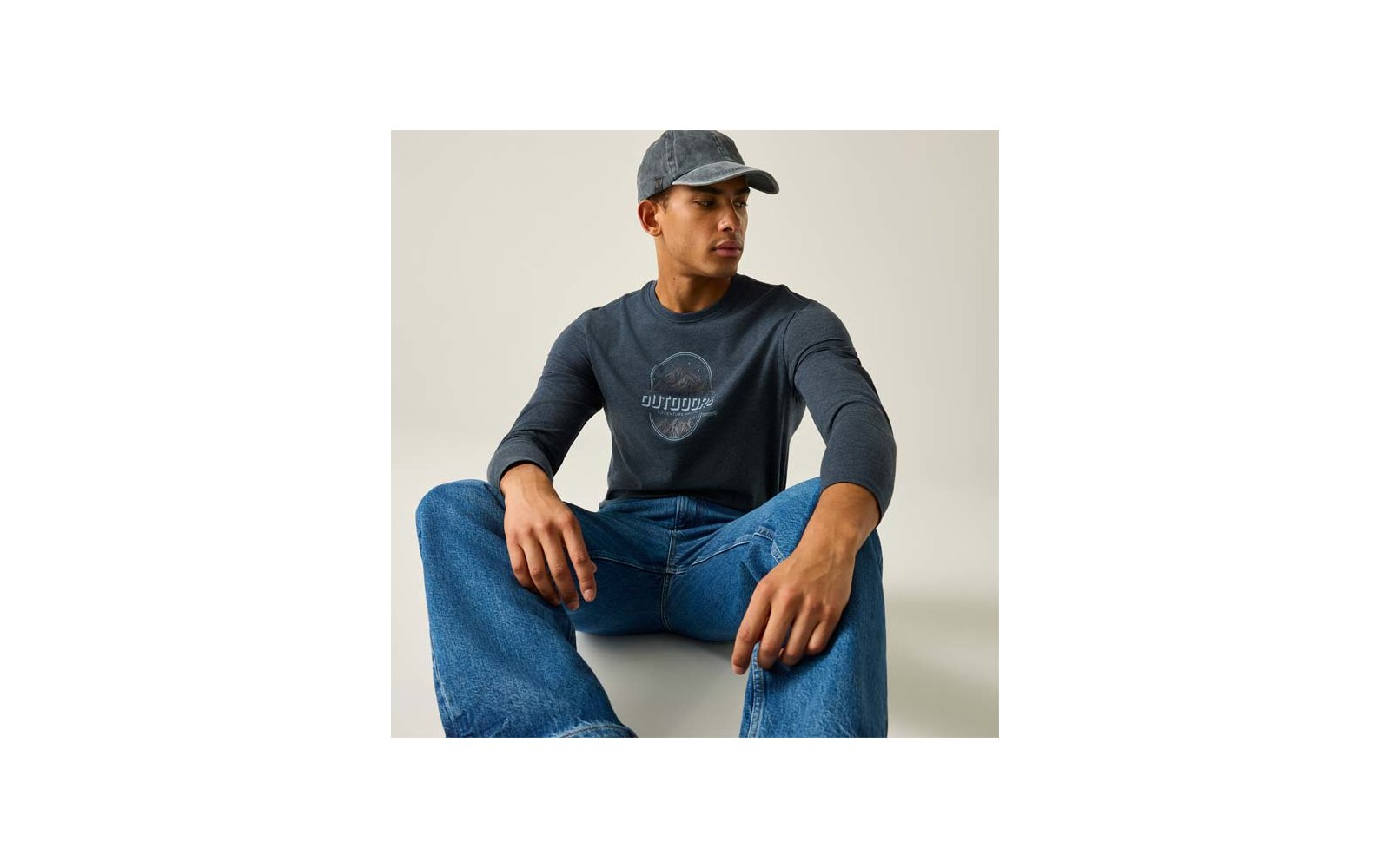 Persoon zit op de vloer, draagt een grijze baseballpet, donker longsleeve met ronde opdruk en blauwe spijkerbroek, kijkt naar rechts