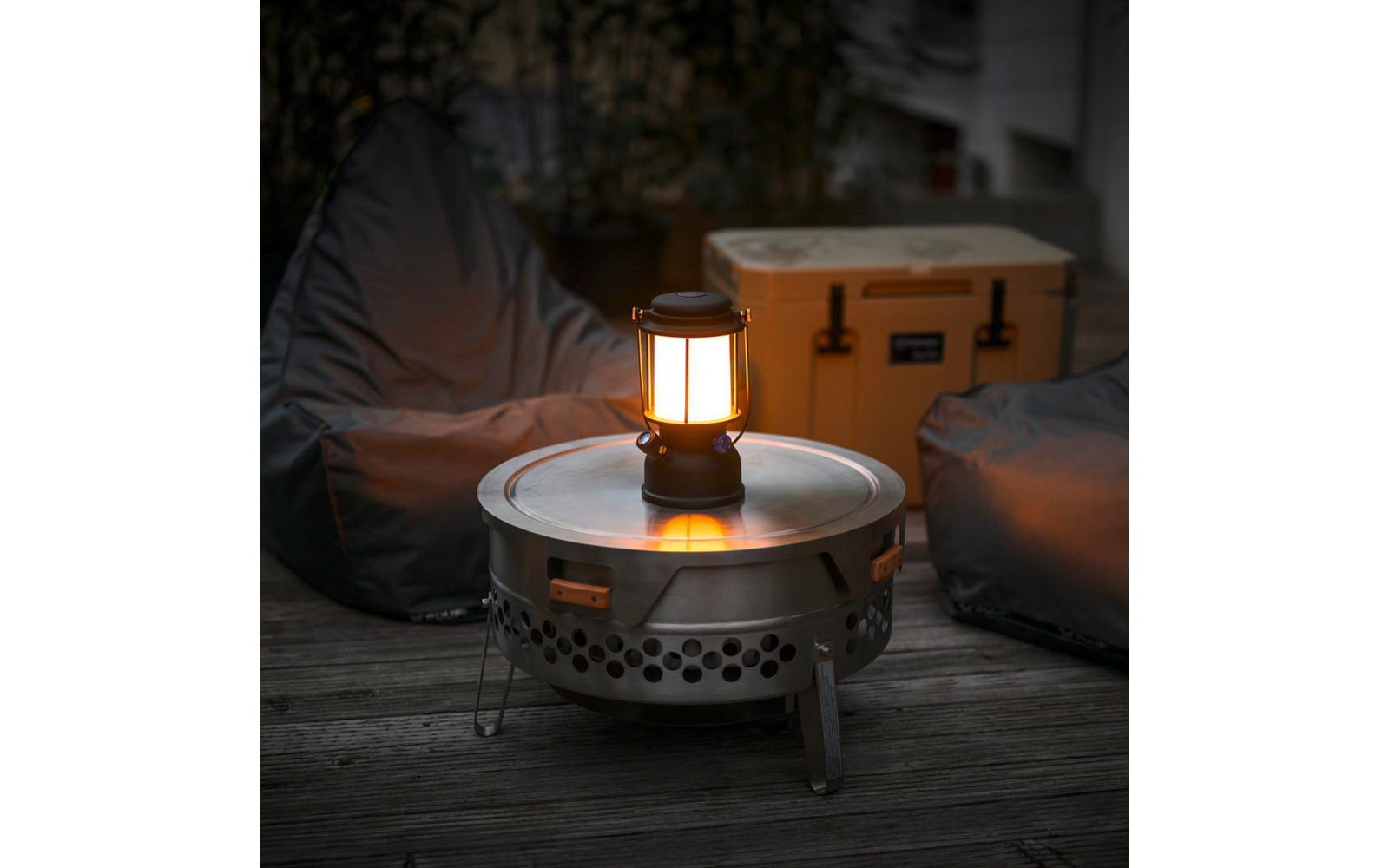 Beleuchtete Laterne auf rundem Metalltisch auf Holzdeck, zwei Sitzsäcke und gelbe Transportbox im Hintergrund