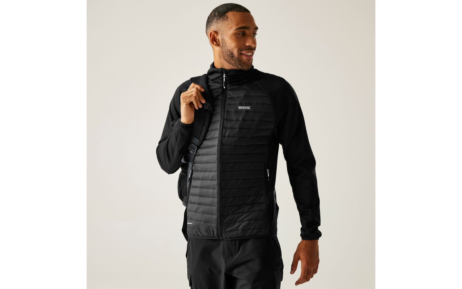 Persoon draagt zwarte doorgestikte jas en zwarte broek, rugzakriem over de rechter schouder, kijkt naar rechts