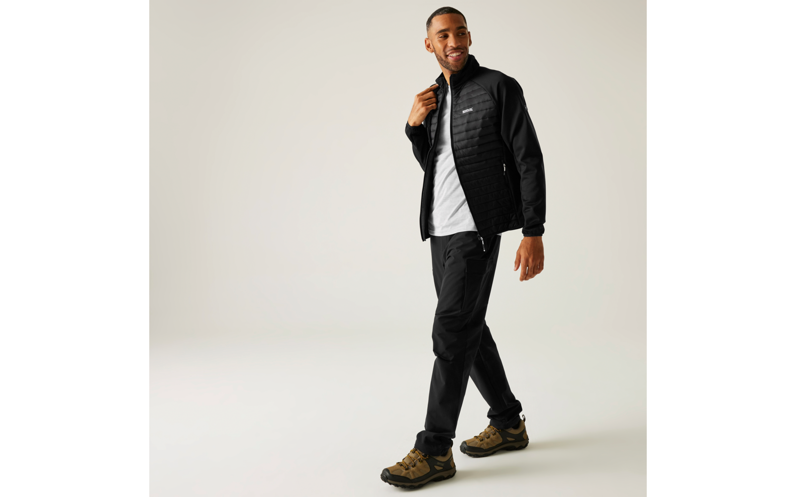 Een man met een zwarte doorgestikte jas, wit T‑shirt, zwarte broek en bruine wandelschoenen voor een lichte achtergrond