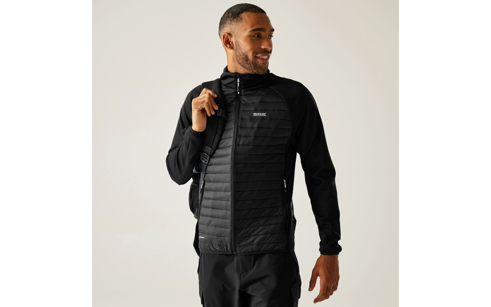 Mann trägt schwarze gesteppte Jacke und schwarze Hose, steht vor heller Wand und lächelt