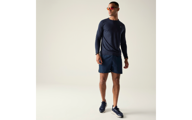 Mann in Ganzkörperansicht vor hellem Hintergrund, trägt dunkelblaues langärmeliges Shirt, dunkelblaue Shorts, dunkelblaue Sportschuhe, orangefarbene Sonnenbrille und Armbanduhr