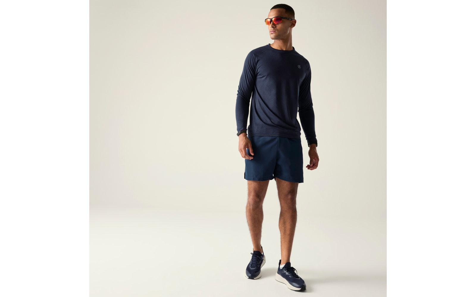 Mann in Ganzkörperansicht vor hellem Hintergrund, trägt dunkelblaues langärmeliges Shirt, dunkelblaue Shorts, dunkelblaue Sportschuhe, orangefarbene Sonnenbrille und Armbanduhr