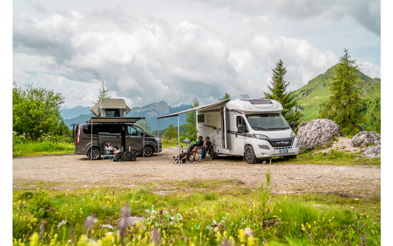 Wohnmobil und Camper mit ausgefahrener Markise auf einem Schotterplatz; zwei Personen sitzen auf Campingstühlen, Berge, Bäume und wolkiger Himmel im Hintergrund