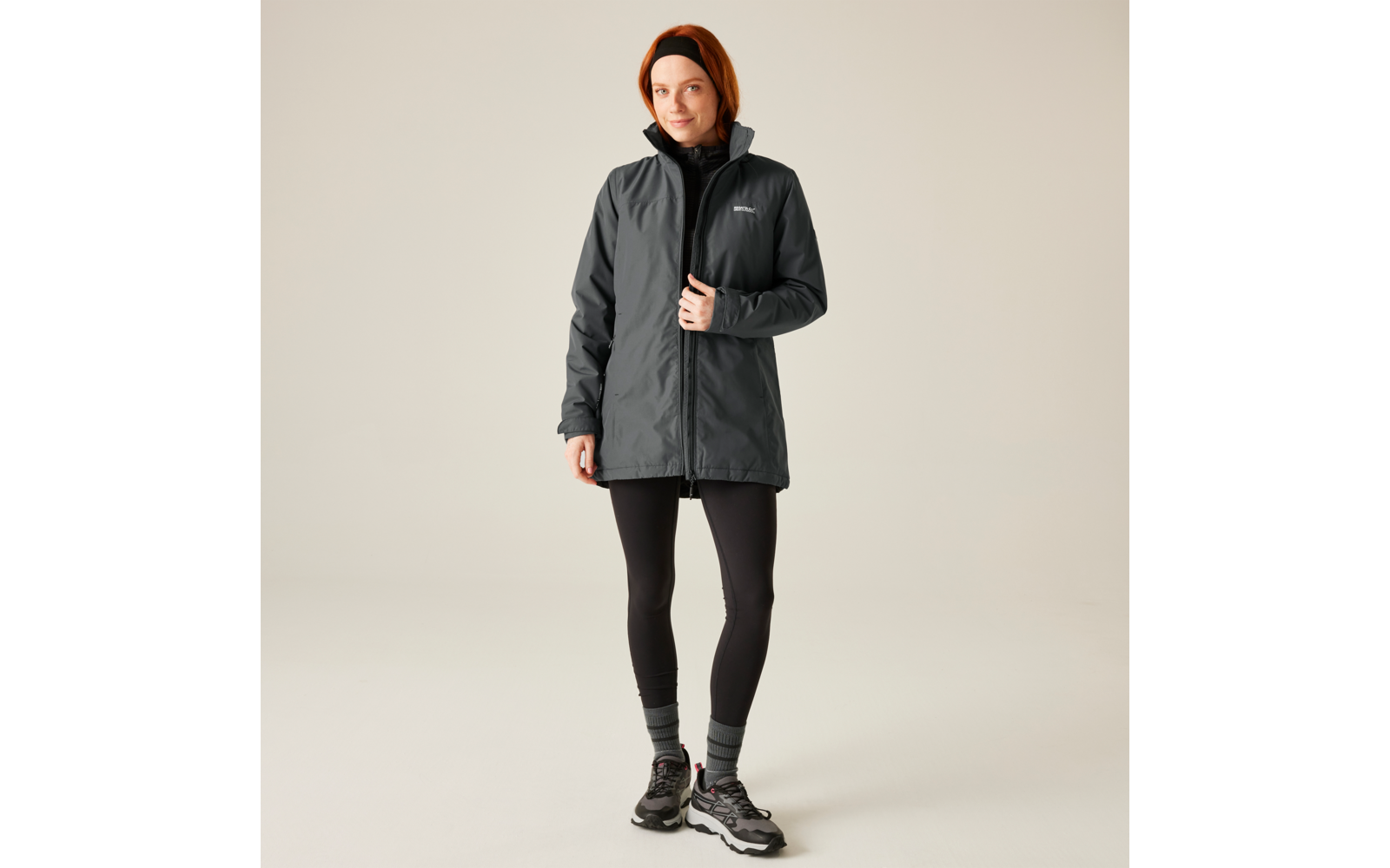 Femme portant une veste grise, des leggings noirs et des chaussures noires, debout devant un fond beige