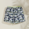 Kurze Shorts mit grau-blauem Camouflage-Muster, neon-grüner Taillenbund und Kordel, auf hellem beigen Hintergrund mit Schatten eines Palmblatts