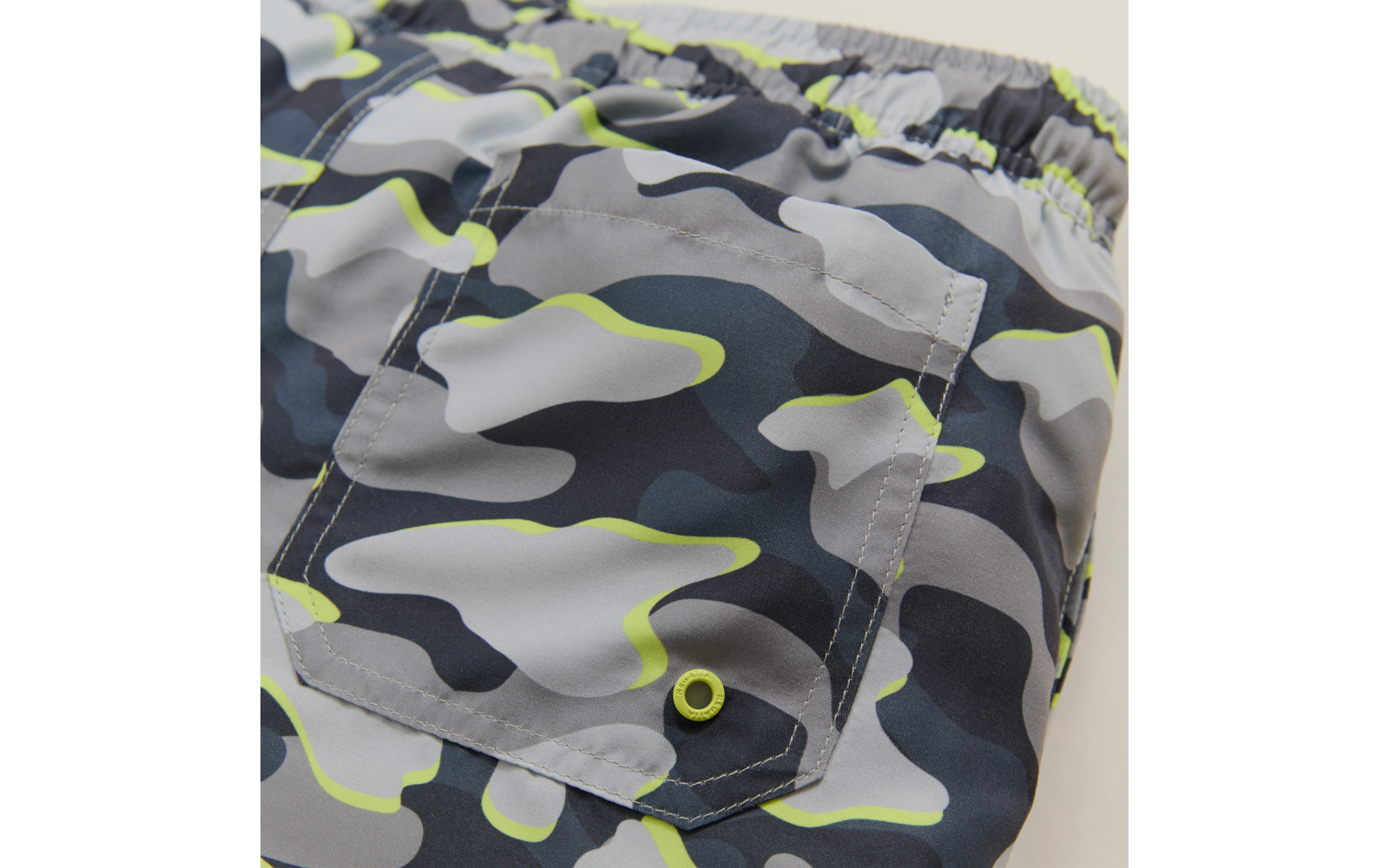 Nahaufnahme eines elastischen Bundes und einer aufgesetzten Tasche aus Stoff mit grau-schwarzem Camouflage‑Muster und neon‑grünen Akzenten, sichtbare Metall‑Öse