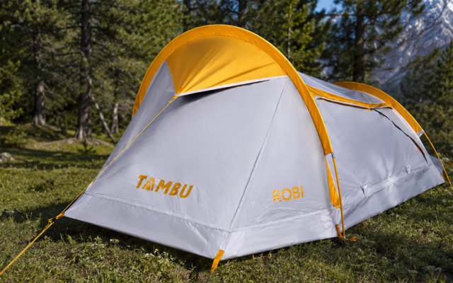 Tienda blanca y amarilla con las palabras 'TAMBU' y 'ROBI' impresas, sobre hierba, con árboles y montañas al fondo